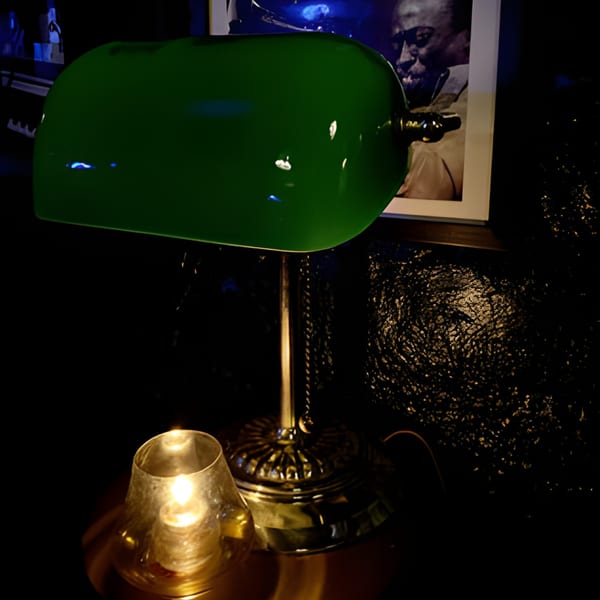 A green banker's lamp and a glowing candle on a dark table, with a framed portrait in the background.