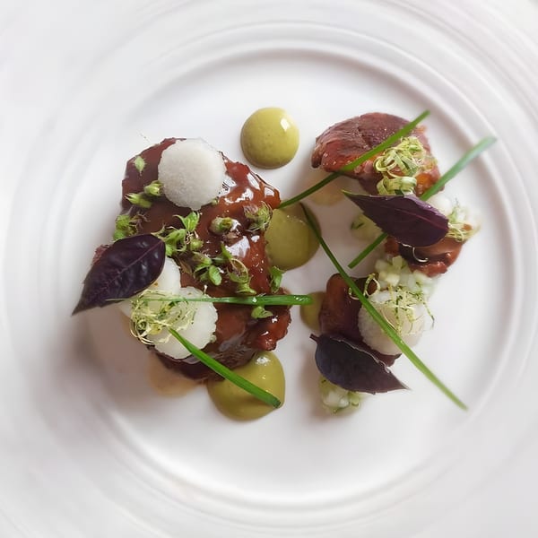 Gourmet steak dish with green sauce, white spheres, purple leaves, and chives on a white plate.