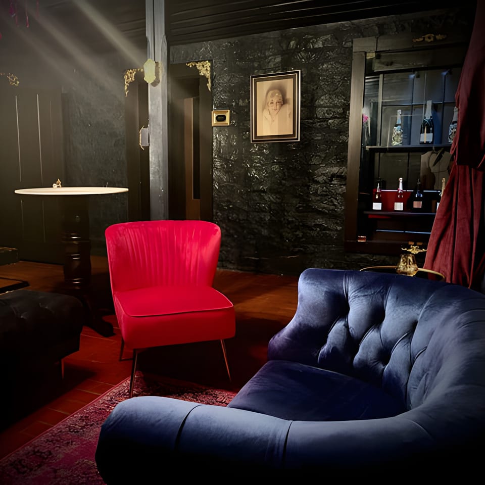 Dark, moody lounge area with red and blue velvet chairs, a small table, and a bar shelf.