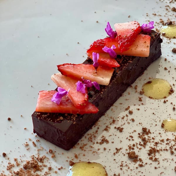 A rectangular chocolate dessert topped with sliced strawberries and purple edible flowers on a white plate.