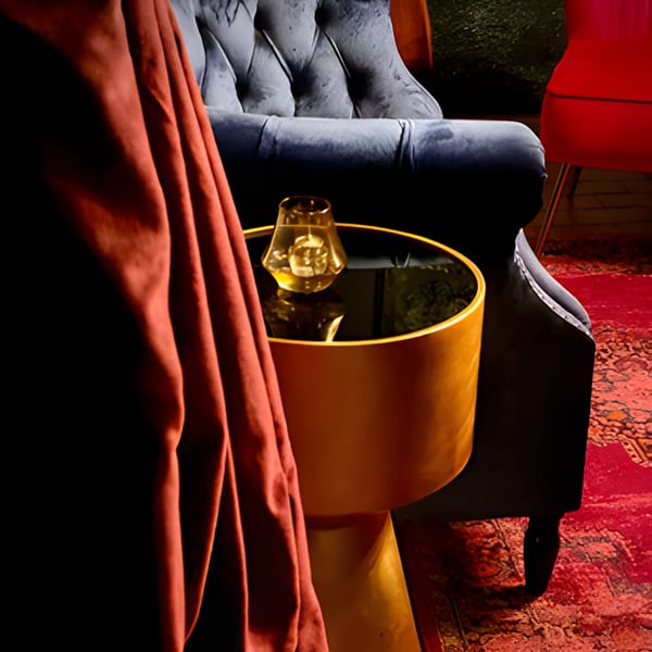 A dark, luxurious interior with a blue velvet armchair, a gold side table, and a red rug.