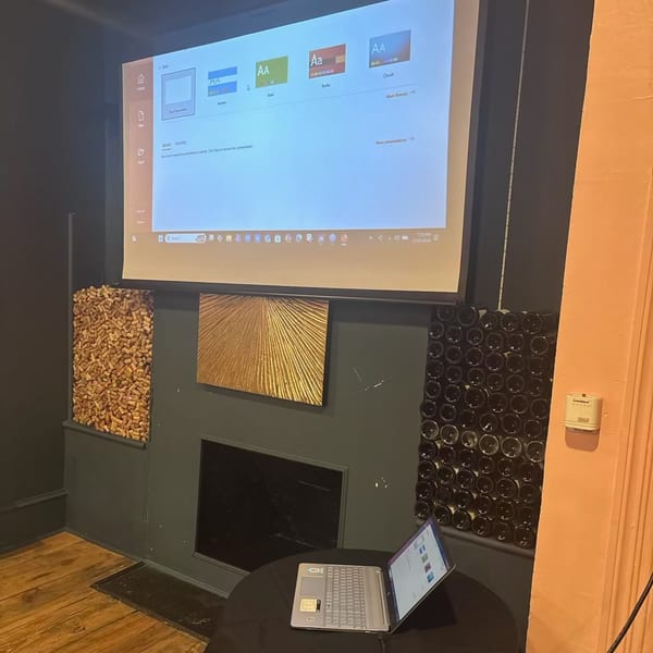A laptop on a table in front of a large screen displaying a presentation, flanked by decorative wine bottles.