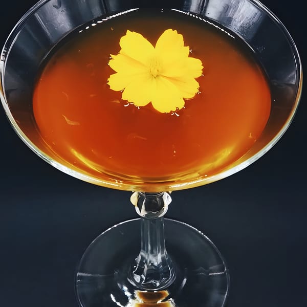 A vibrant yellow flower floats atop a rich amber cocktail in a delicate martini glass.