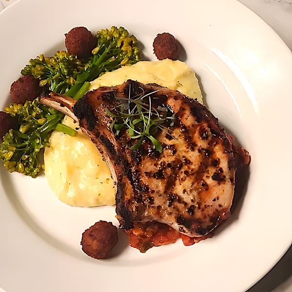 Grilled pork chop with mashed potatoes, broccolini, and small fried balls on a white plate.