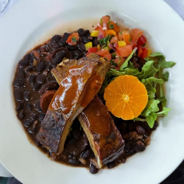 Two ribs with sauce, black beans, pico de gallo, greens, and an orange slice on a white plate.
