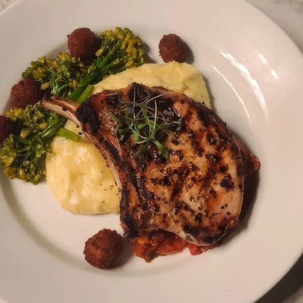 Grilled pork chop with mashed potatoes, broccolini, and hush puppies on a white plate.