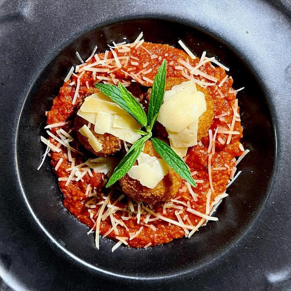 Fried arancini balls with tomato sauce, shredded and shaved parmesan, and fresh mint in a black bowl.