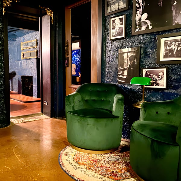 Two green velvet armchairs on a patterned rug, with a dark textured wall adorned with framed photos.