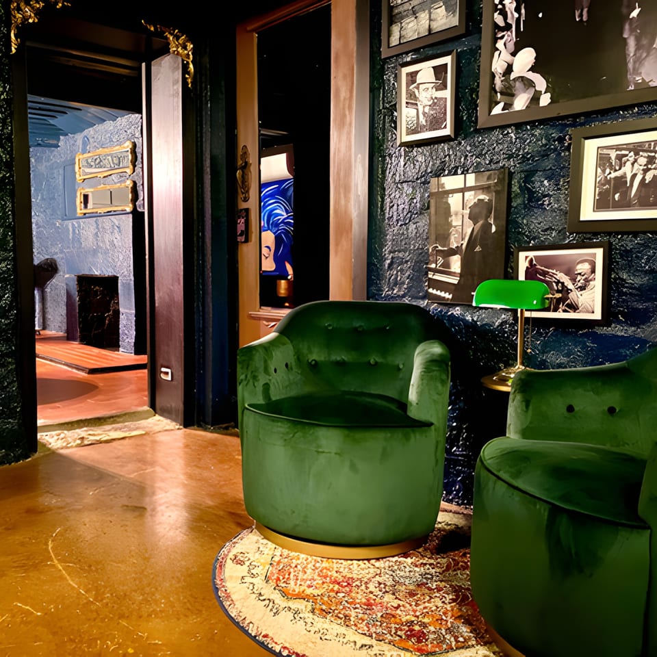 Two green velvet armchairs on a patterned rug, with a dark textured wall adorned with framed photos.